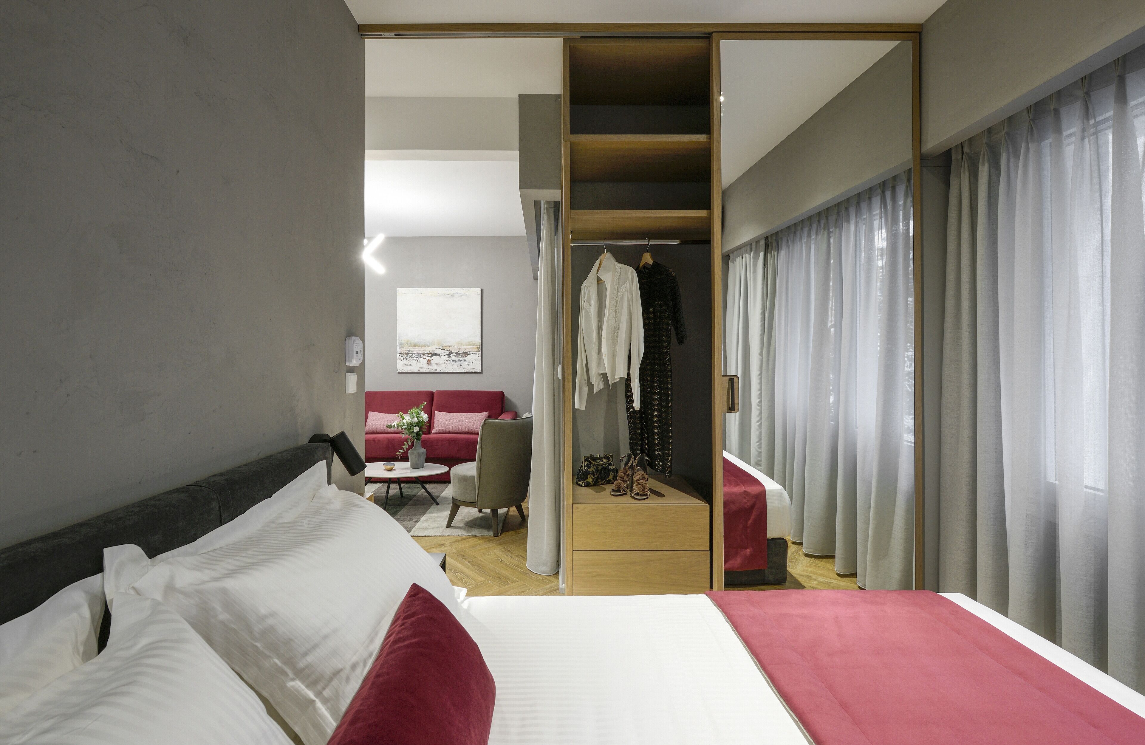 Una habitación de hotel moderna con una cama grande, un sofá rojo, una mesita con una planta y un vestidor con ropa colgada.