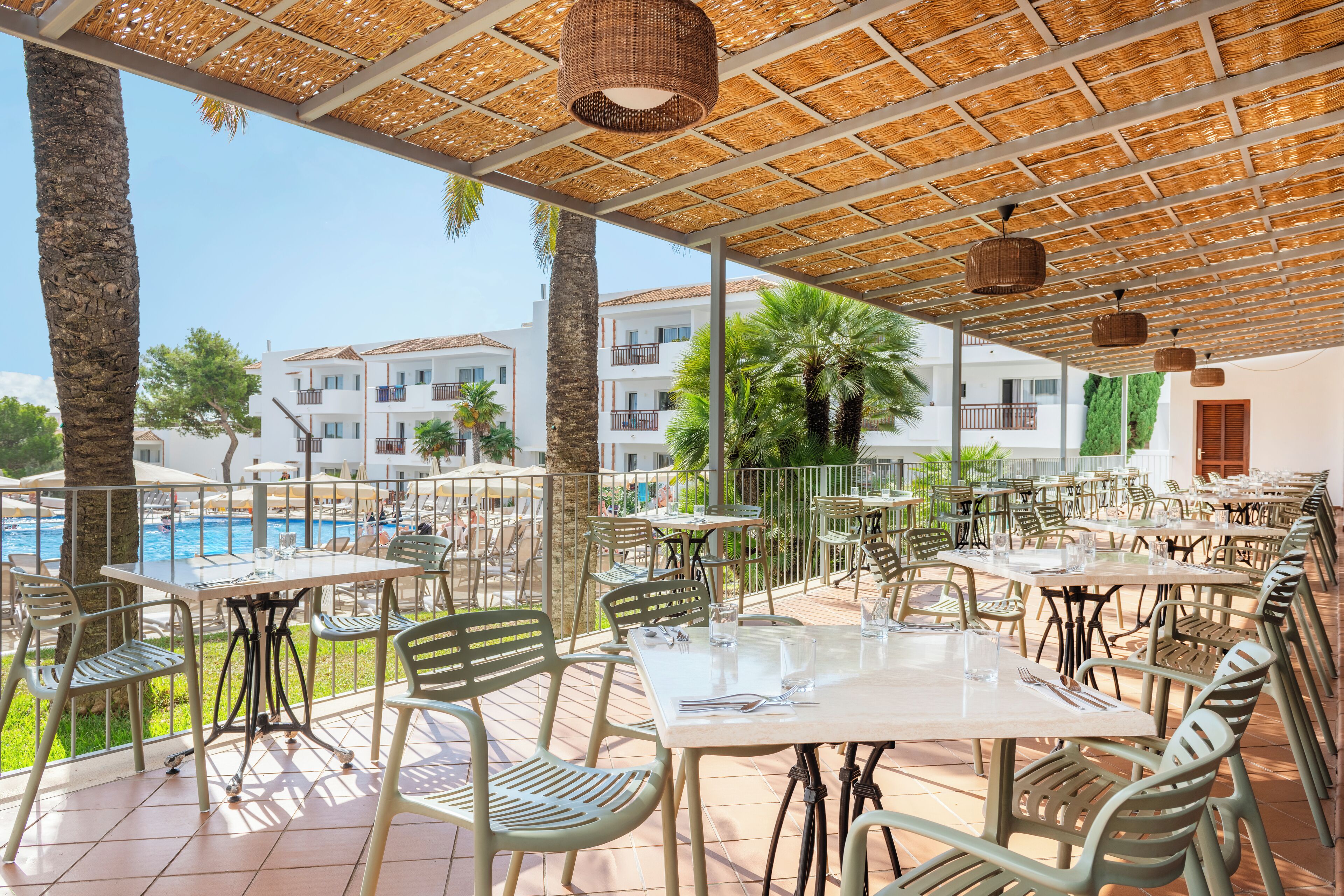 A covered outdoor dining area with tables and chairs, overlooking a pool and buildings.