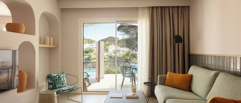 A modern living room with a sofa, a coffee table, a TV, and a view of a pool and greenery.