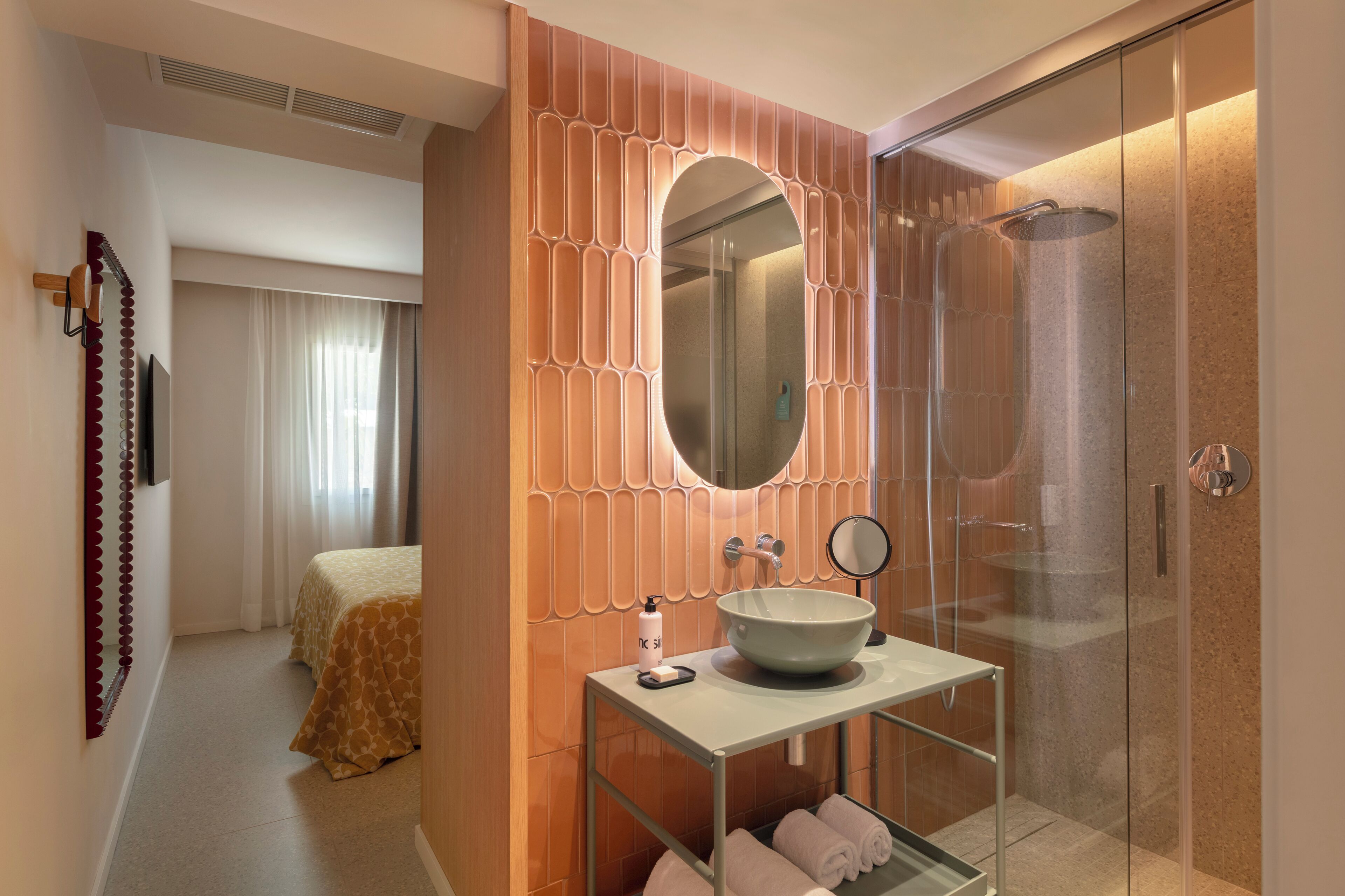 A modern bathroom with a large oval mirror, a vessel sink, a glass shower enclosure, and a bed with a patterned bedspread.