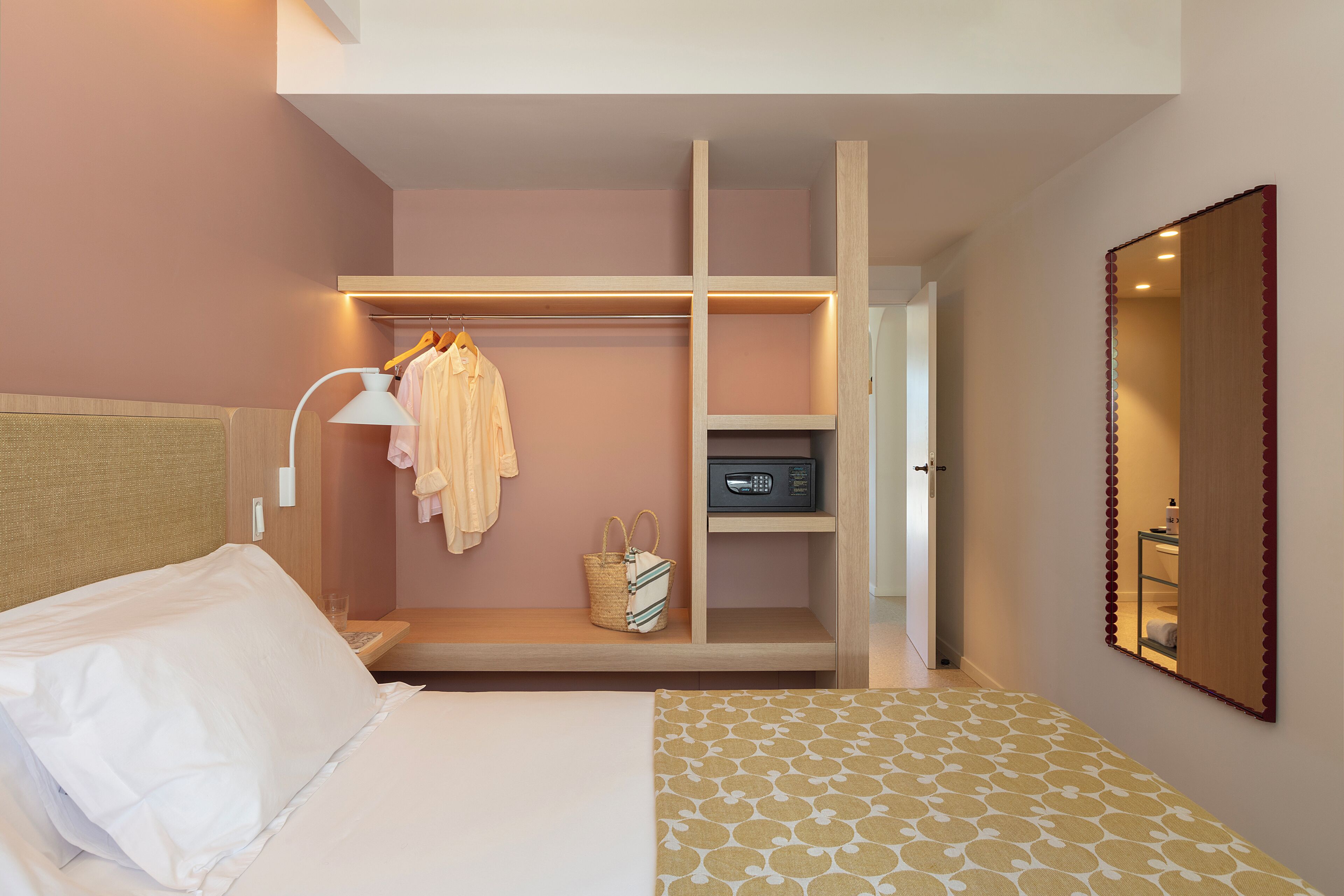 A bedroom with a bed, a bedside table with a lamp, a wardrobe, a mirror, and a shelf with a radio.
