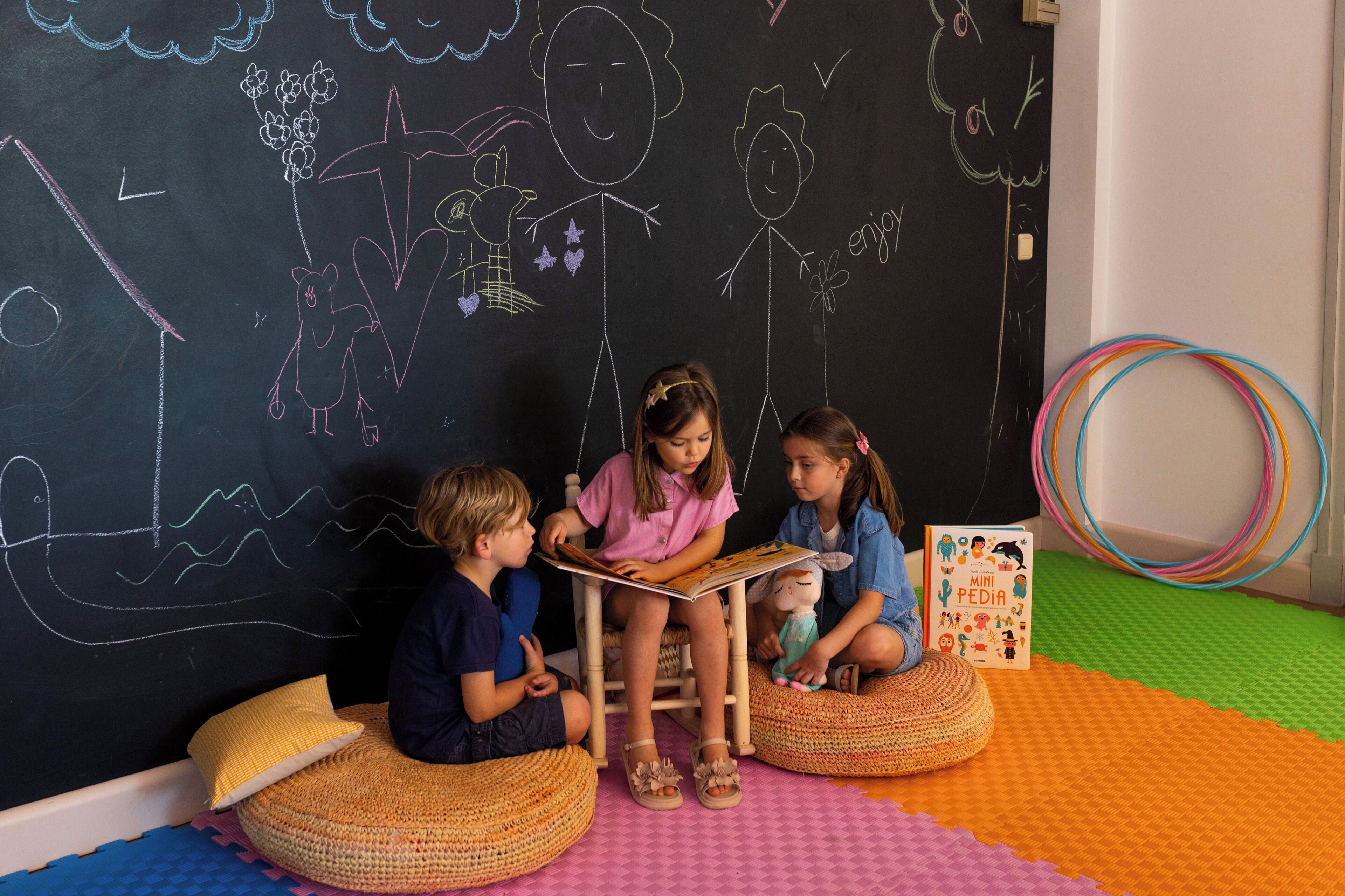 Children sitting on woven floor cushions in front of a blackboard with drawings.