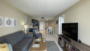A living room with a sofa, coffee table, and TV. There is a dining area with a table and chairs. The kitchen is visible in the background. - BLDG Construction Aug-Oct 2026; See Desc below! Sea Winds 1203 (Marco Island)