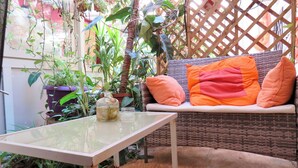 Um sofá de vime com almofadas laranja, uma mesa com tampo de vidro e diversos vasos com plantas.