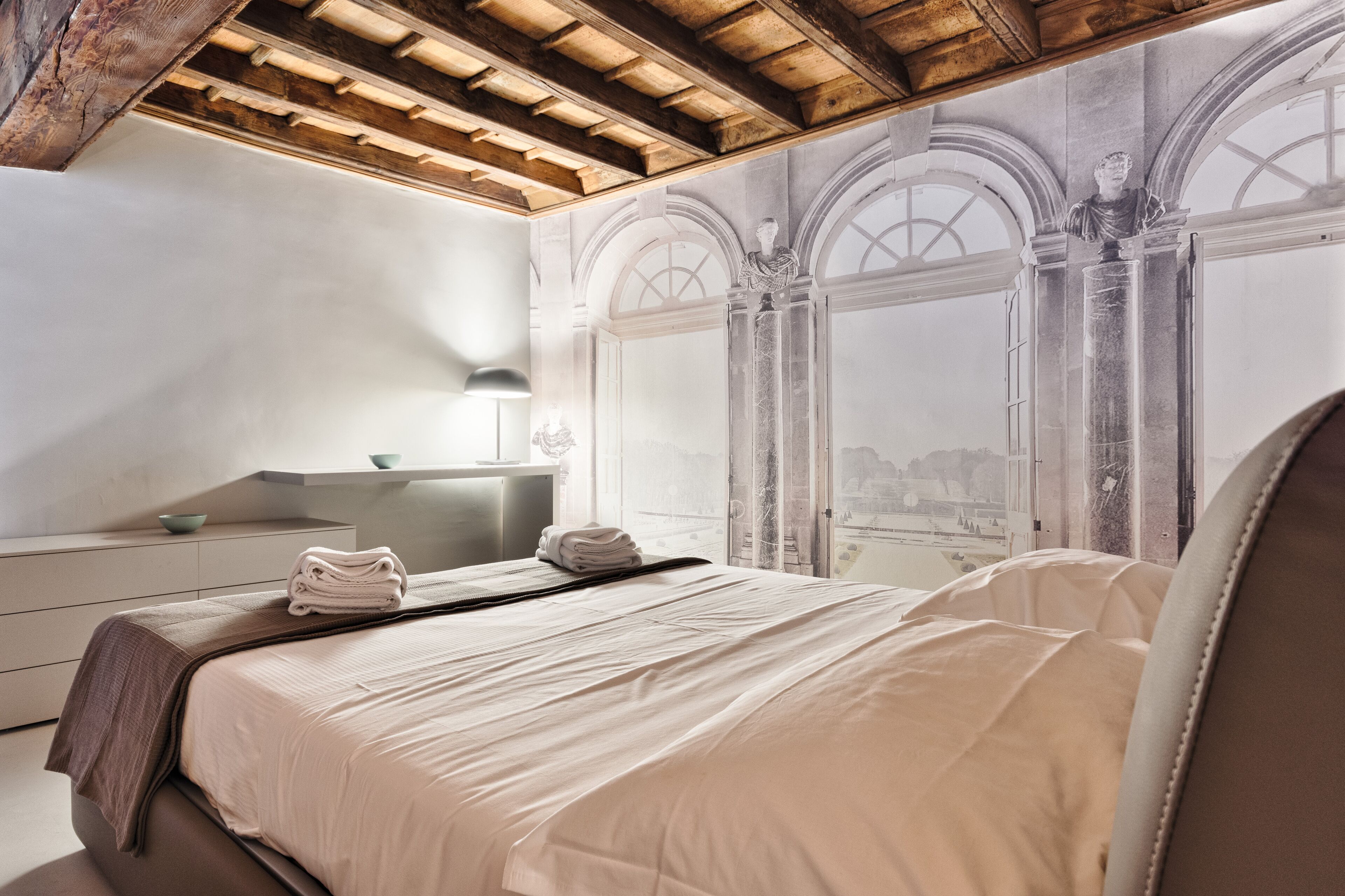 Habitación de hotel moderna con una cama grande, una mesita de noche, una lámpara y vistas a la ciudad a través de ventanales abovedados.