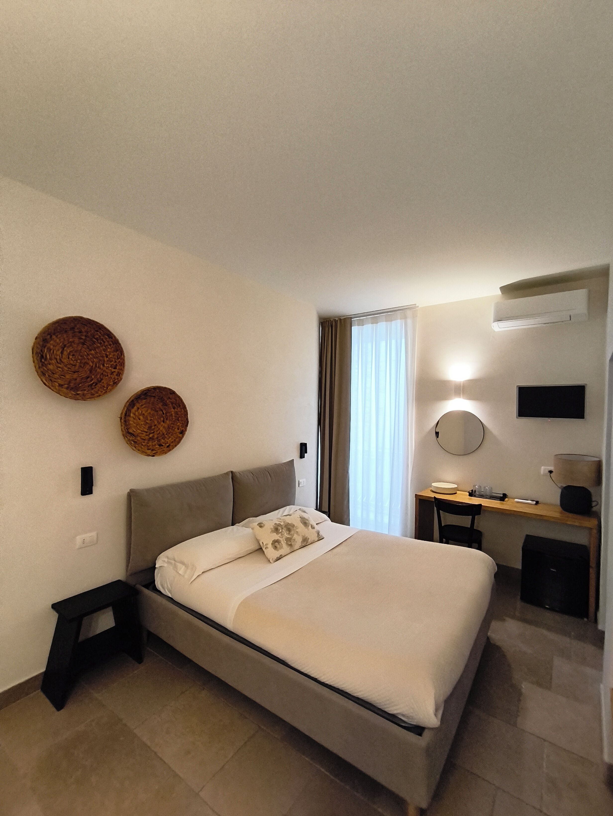 Habitació d'hotel amb un llit, una taula de treball, una cadira, un mirall i un aire condicionat fixat a la paret.