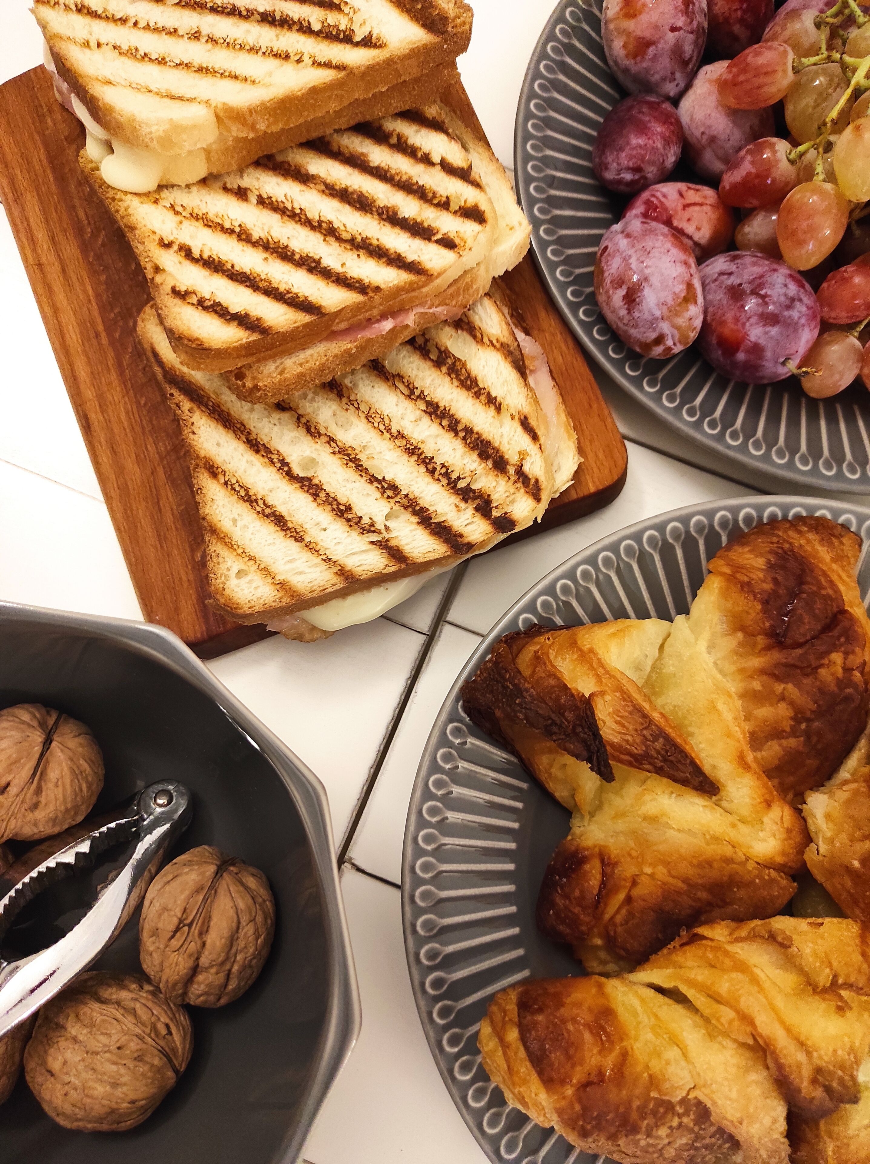 A wooden cutting board with grilled sandwiches, a plate of grapes, a bowl of walnuts, and a plate of pastries.