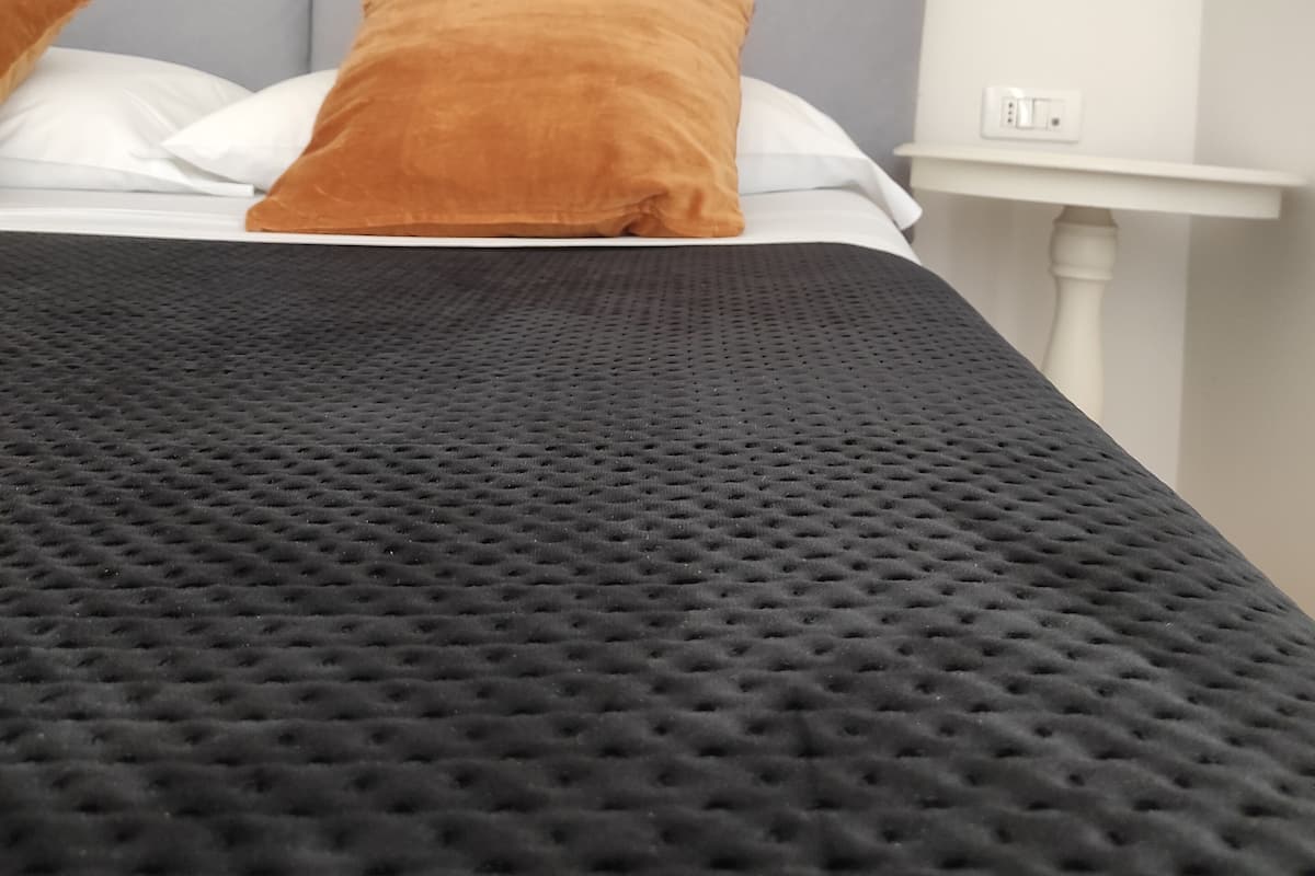 A bed with a textured black bedspread, two pillows, and a woven basket on the wall.