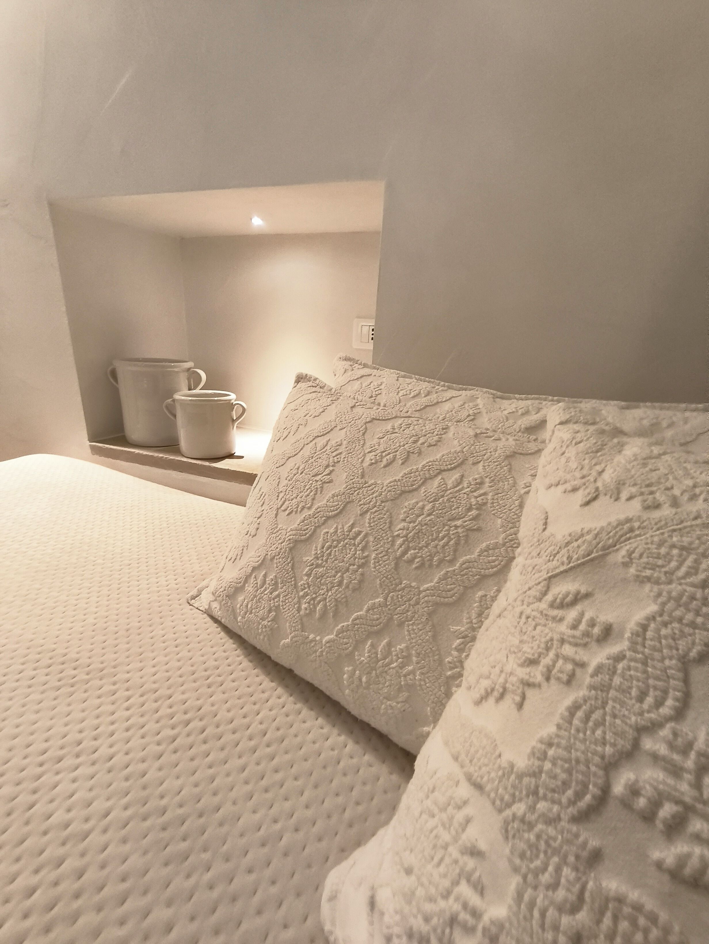 A bed with a textured white cover and a decorative pillow, a small shelf with two white mugs, and a wall socket.