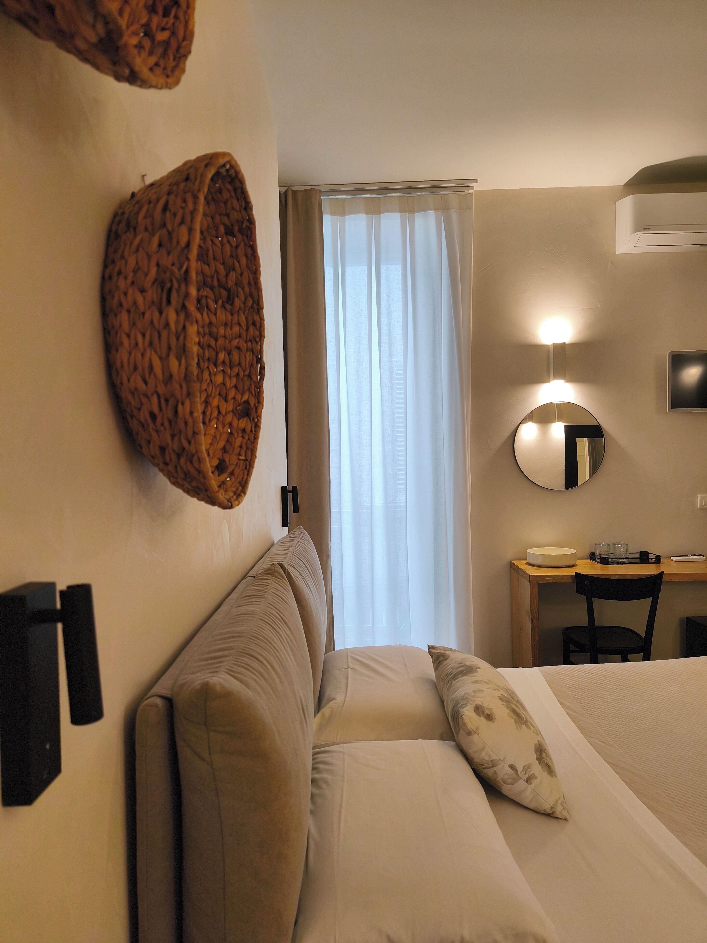Una habitación de hotel con cama, escritorio, silla, espejo redondo y lámpara de pared.