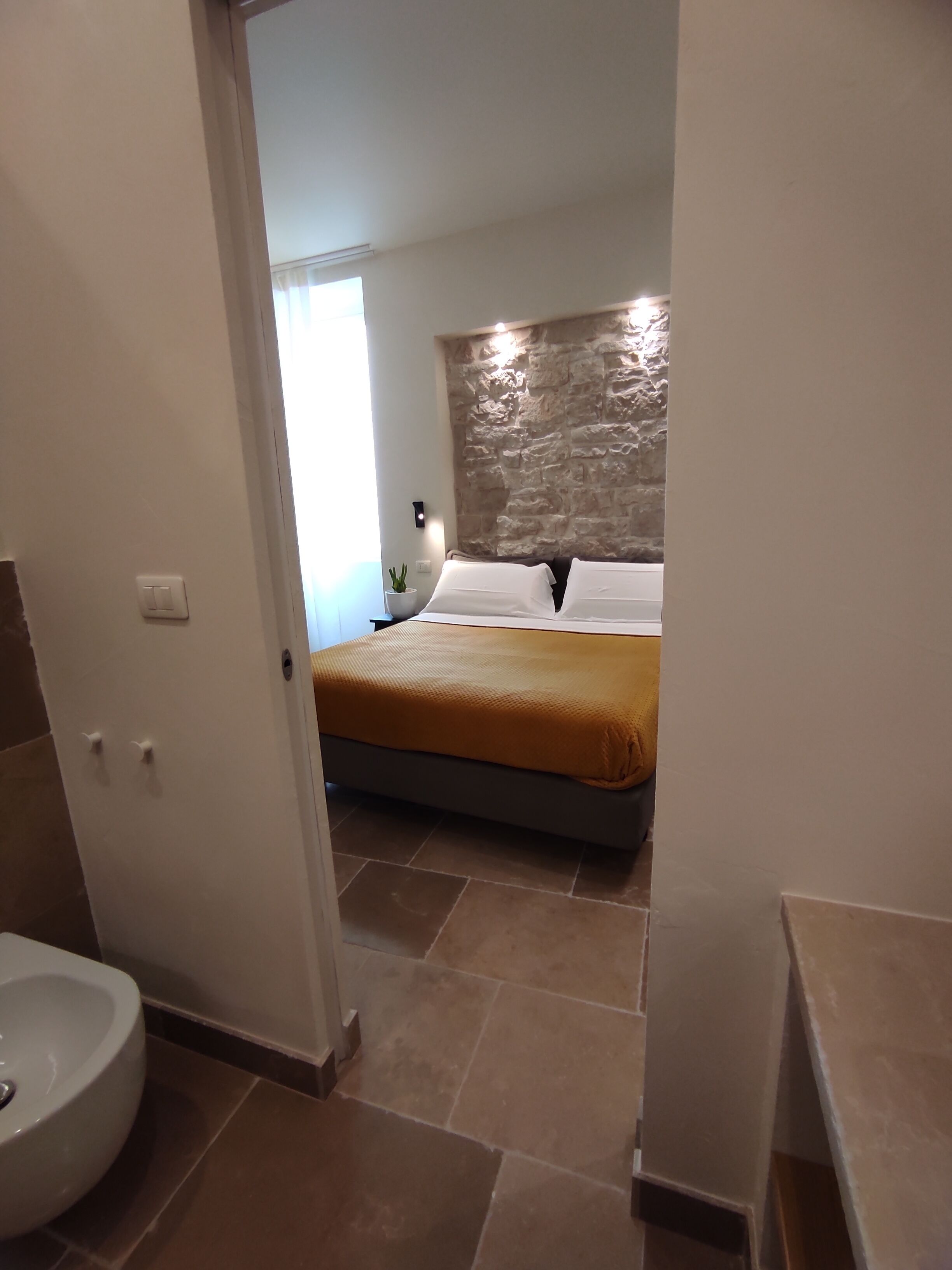 Ein Schlafzimmer mit einer Wand aus Stein, einem Bett mit weißen Leinen und einem kleinen Nachttisch mit einer Lampe.