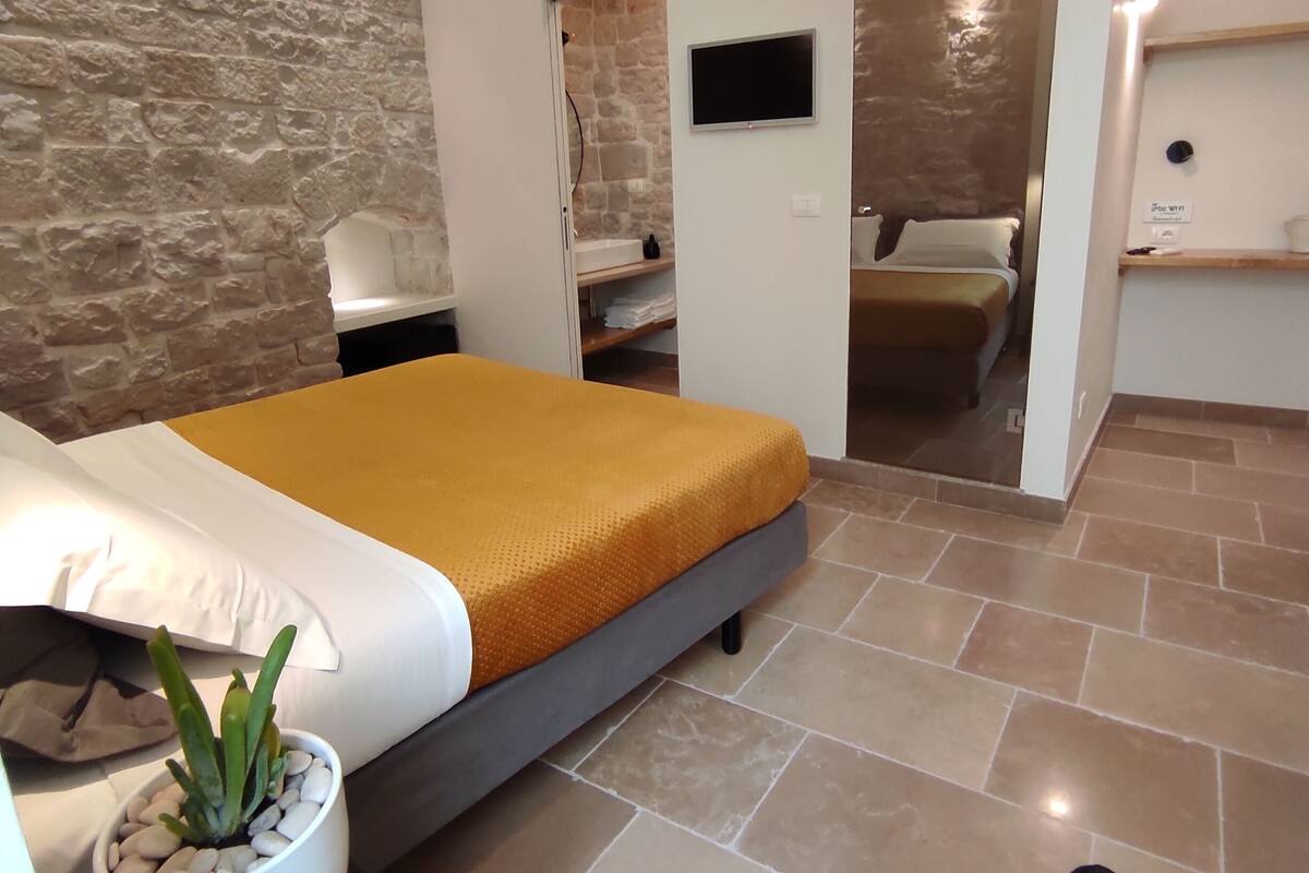 A modern bedroom with a stone accent wall, a bed with a mustard-colored blanket, a small potted plant, and a flat-screen TV mounted on the wall.