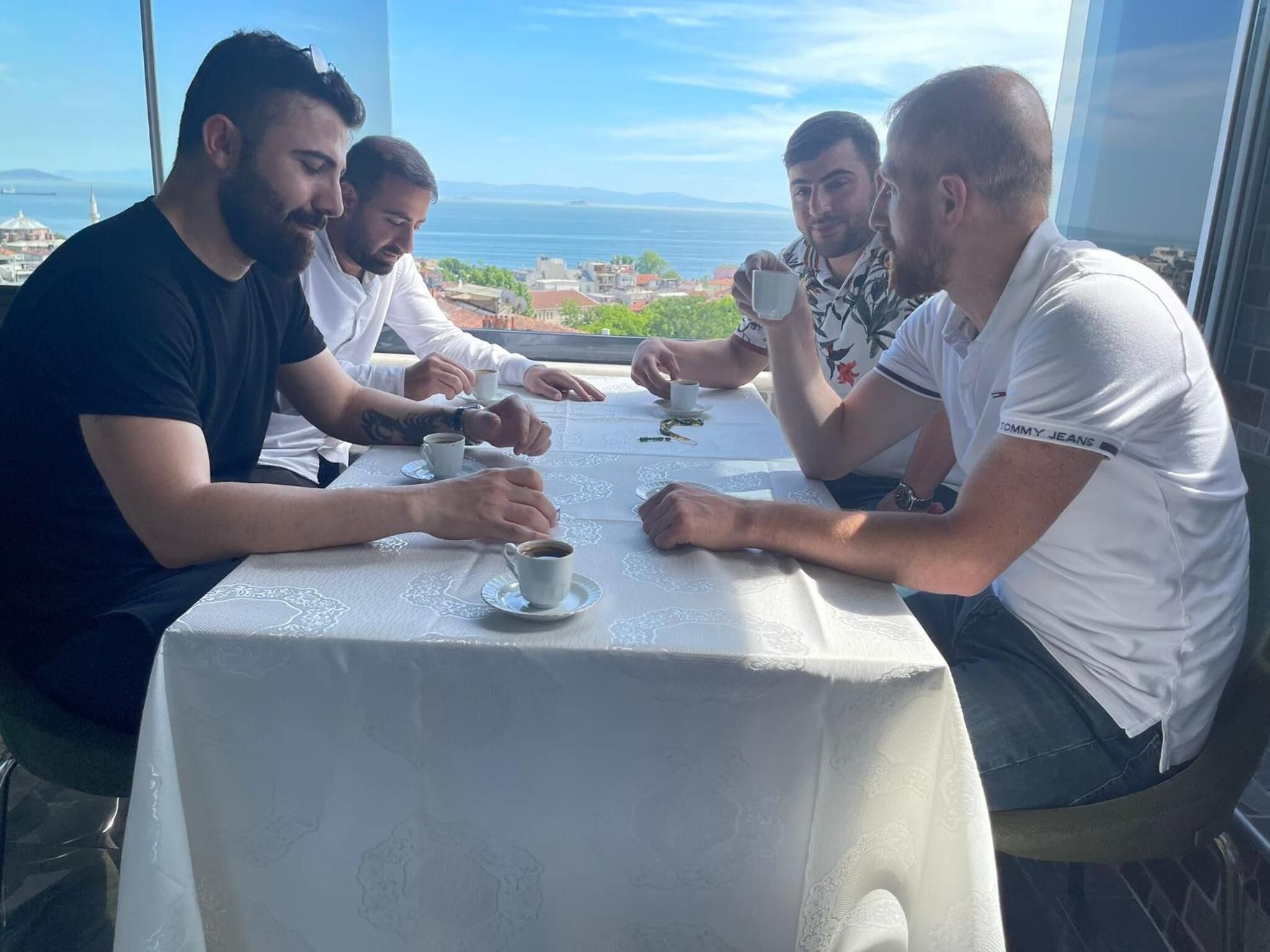 A group of people sitting at a table with a view of the sea and buildings in the background.