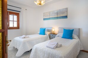 A hotel room with two beds, a painting of a seascape, a chandelier, and a window with curtains.