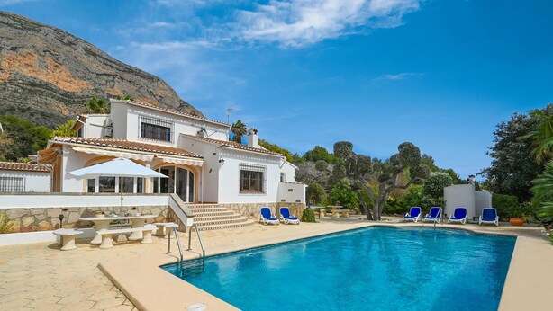 Un espace piscine avec une piscine à l’eau d’un bleu limpide, un bâtiment blanc doté d’un toit en tuiles et des chaises longues bleues.