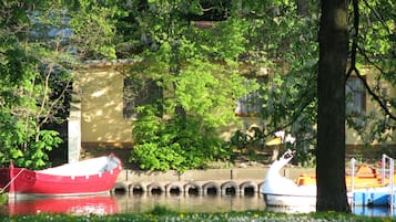 Un bote rojo amarrado en un canal, cerca hay una embarcación con forma de cisne, todo rodeado de vegetación exuberante y un edificio al fondo.