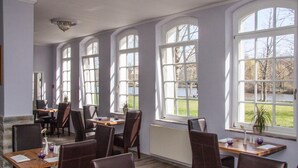 Comedor con varias mesas y sillas, ventanas amplias con vista a árboles y una masa de agua, y una pared de piedra en un lateral.