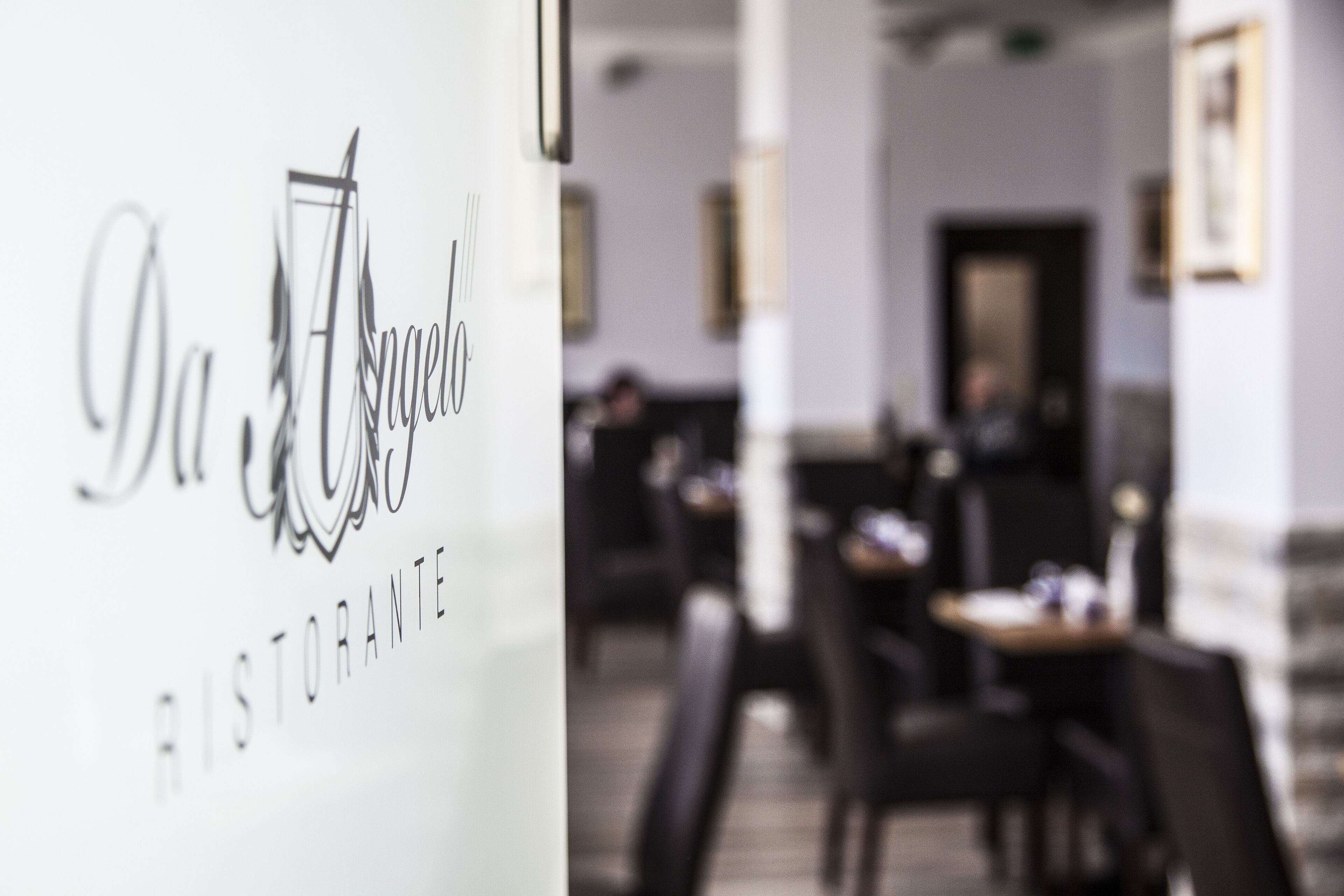 A restaurant interior with a modern design, featuring a white wall with the restaurant's name and logo, and a blurred background of tables and chairs.