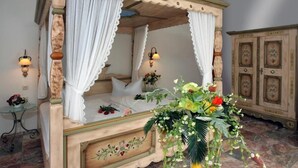 Una cama con dosel y cortinas blancas, un arreglo floral sobre la mesita de noche y un armario de madera al fondo.