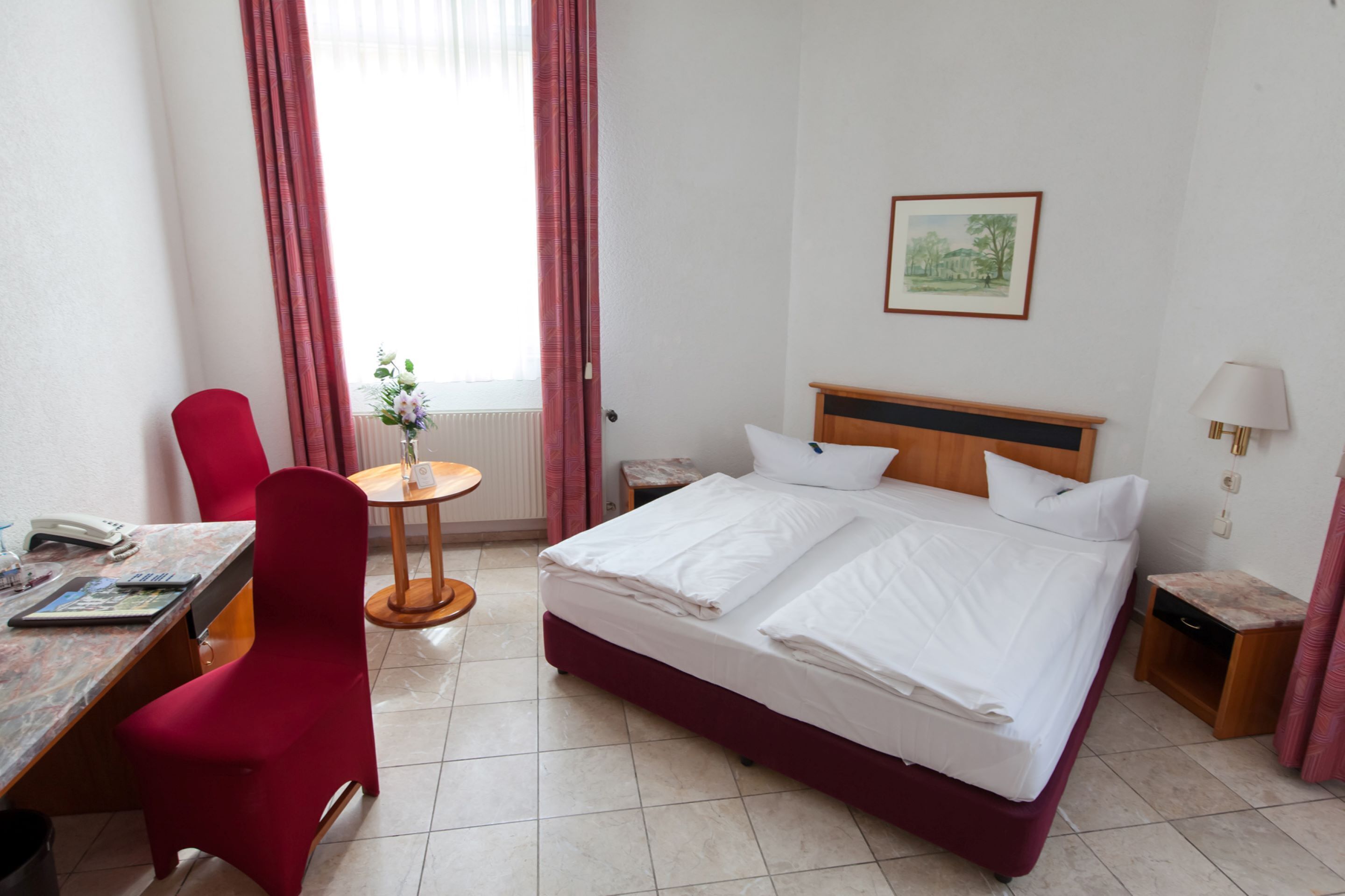A hotel room with a bed, a desk, a chair, a small table with a vase, and a window with curtains.
