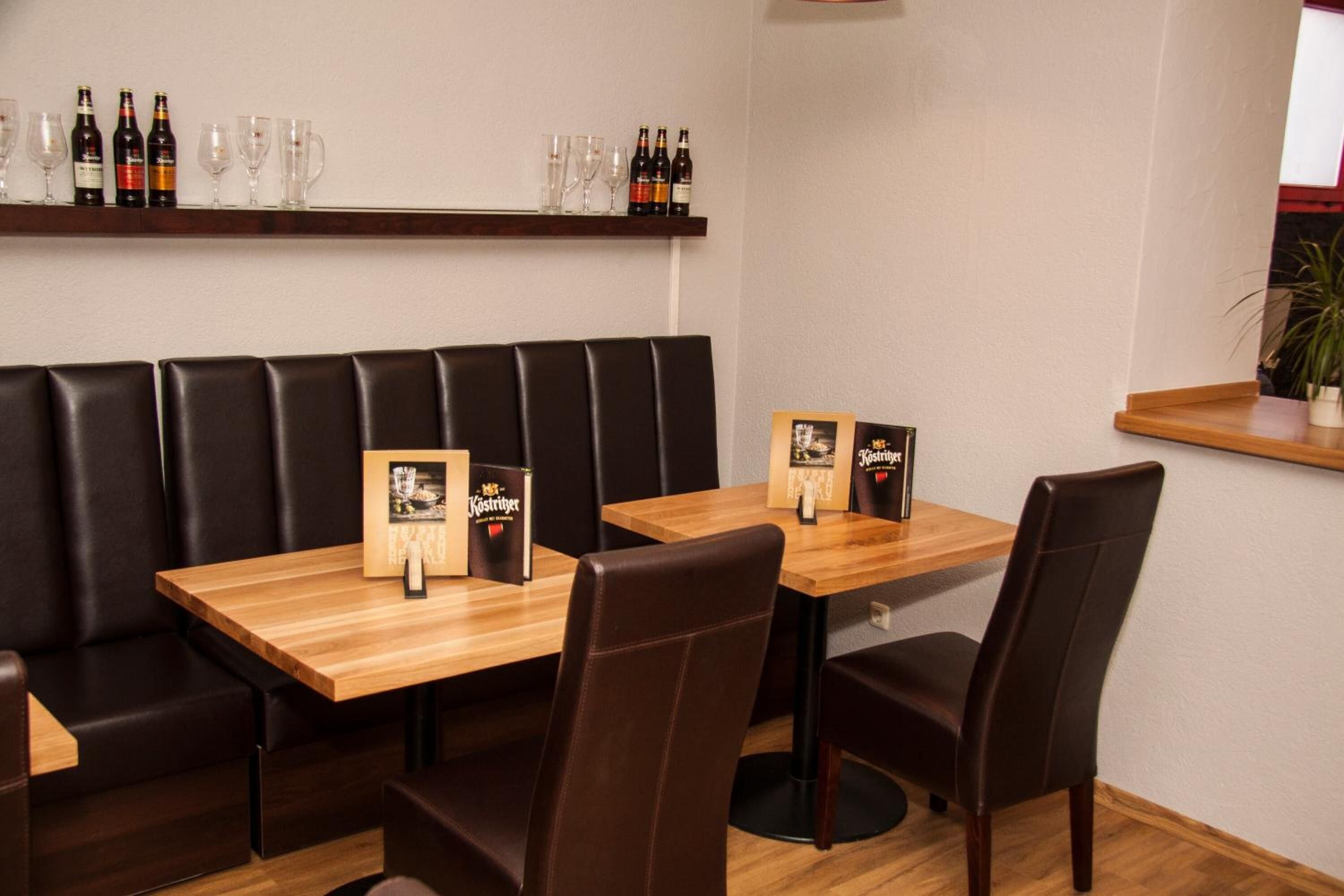 L’intérieur d’un restaurant présente des tables en bois et des banquettes en cuir noir, des étagères remplies de bouteilles et de verres, ainsi que des menus encadrés.