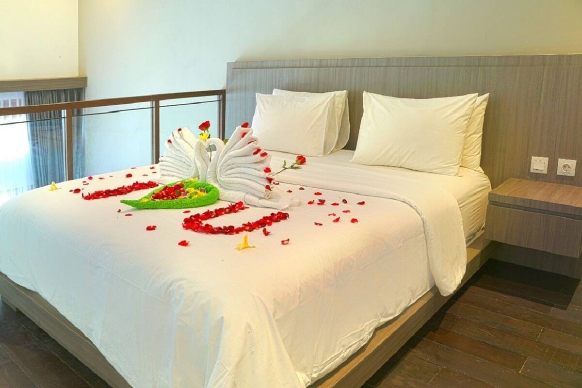 A neatly made bed with white linens, a green floral arrangement, and red rose petals scattered on the bed.