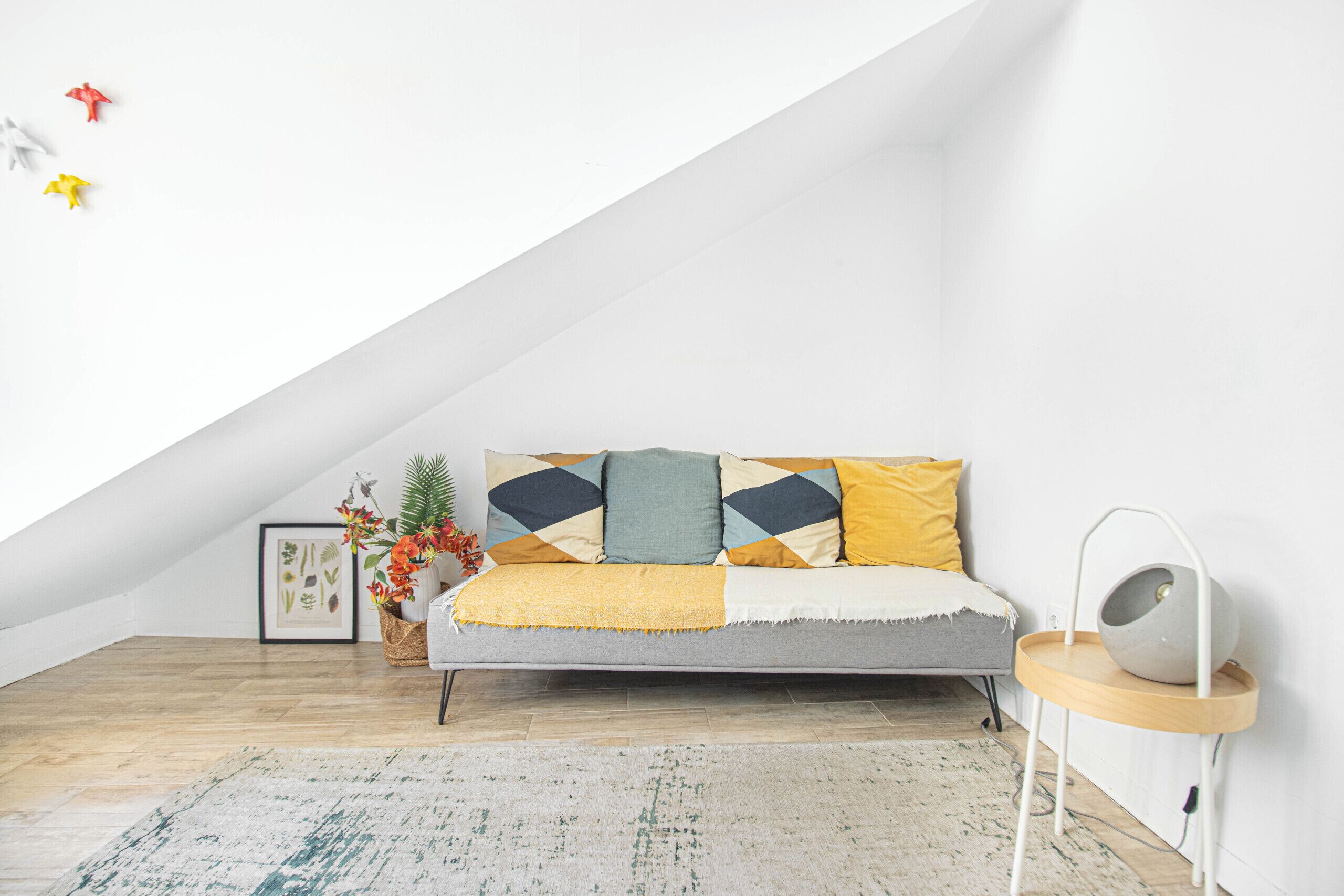 An attic room with a sloped ceiling, a sofa with patterned cushions, a side table with a lamp, and a framed botanical print on the wall.