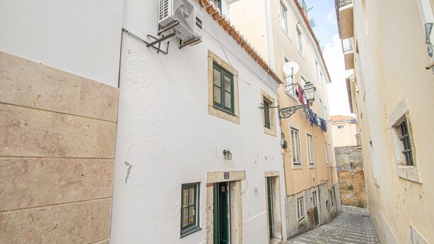 Una estrecha calle empedrada flanqueada por edificios con fachadas blancas y beige, ventanas pequeñas y una puerta visible.