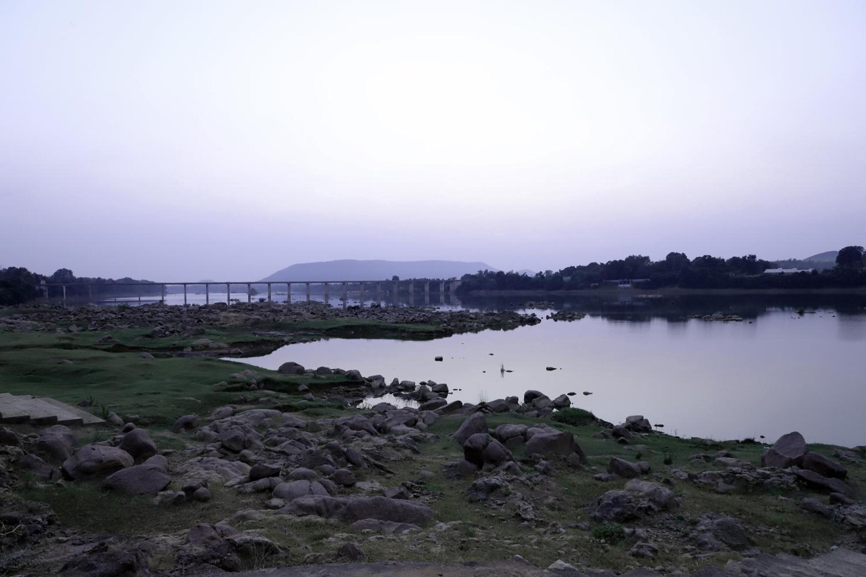 A serene lakeside landscape with a bridge, rocky shore, and distant hills.