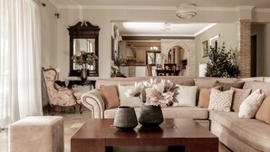 A living room with a sofa, ottoman, and coffee table. There is a chair with a floral pattern and a mirror on the wall. - Amalthia Villa (Zakynthos)