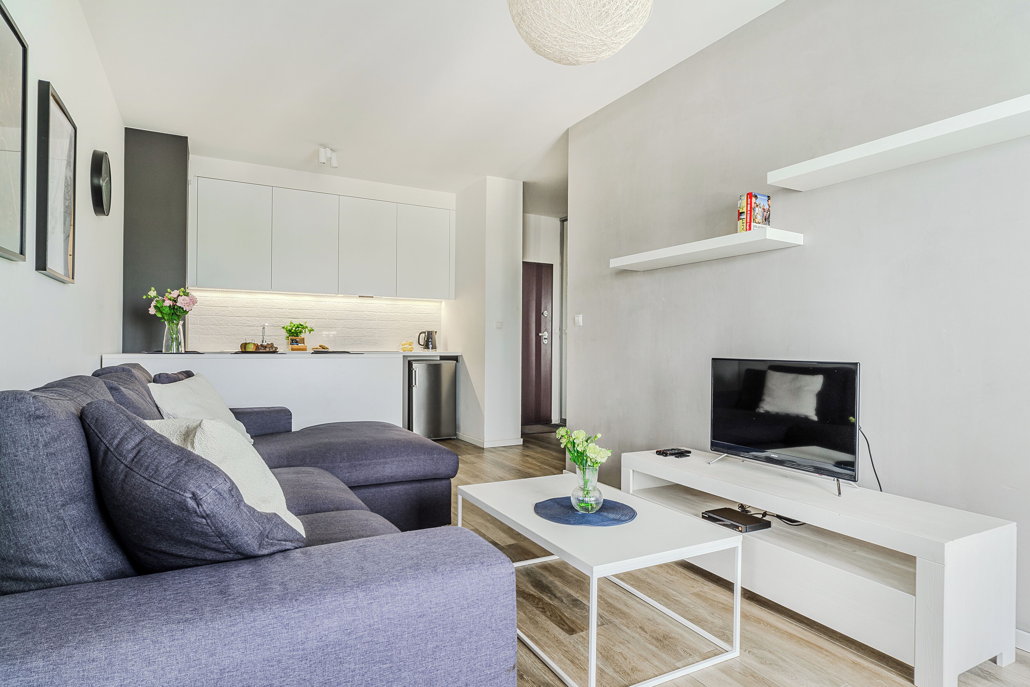 Una sala de estar moderna con un sofá gris, una mesa de centro blanca con un jarrón, un televisor de pantalla plana sobre un soporte blanco, y una zona de cocina al fondo.