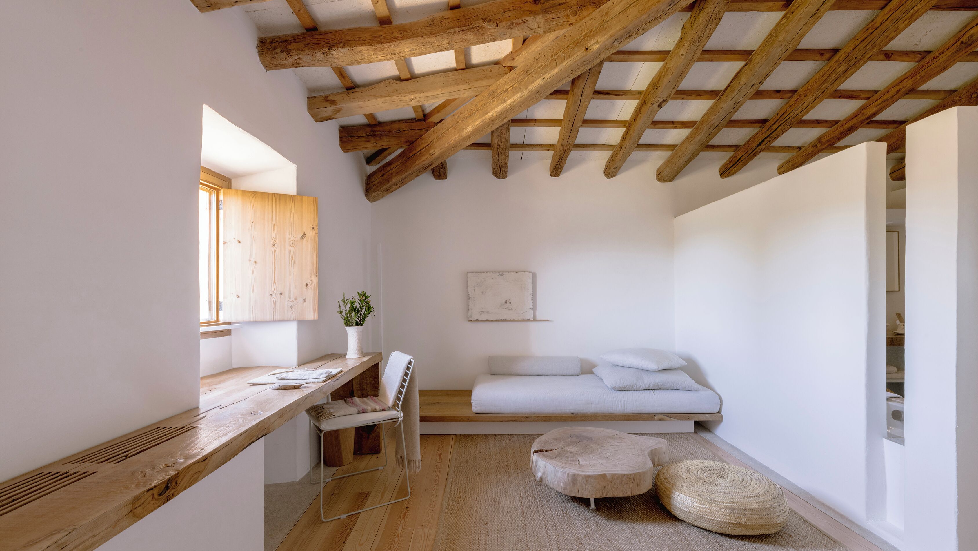 Una habitación pequeña y acogedora con un escritorio de madera, una cama con almohadas y un ventanal grande.