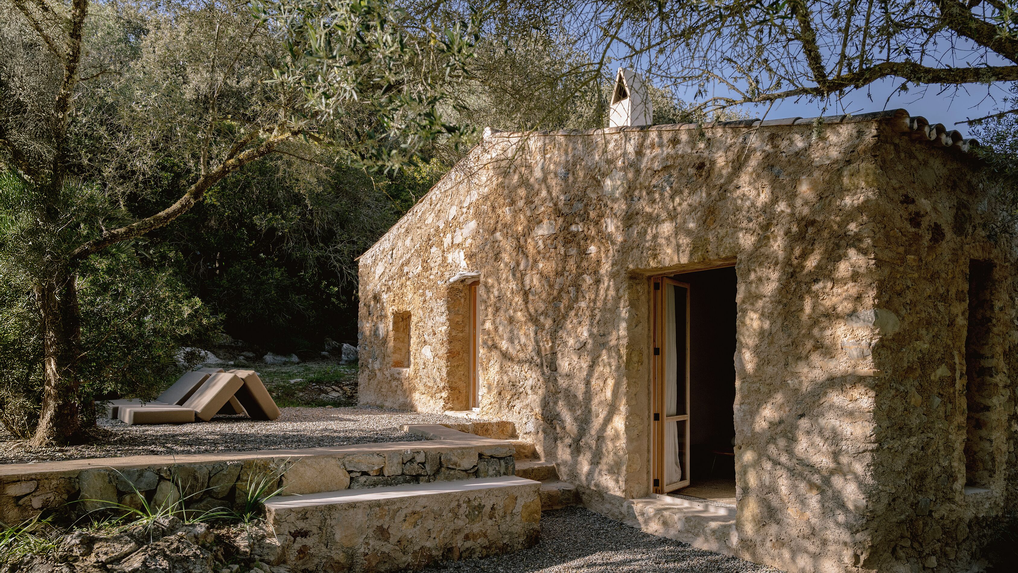 A rustic stone building with a wooden door and window, surrounded by trees and gravel.