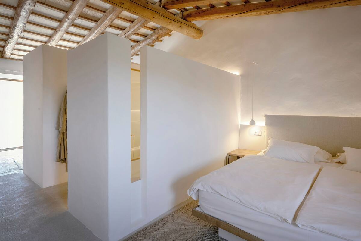 Ein Schlafzimmer mit Holzboden, einem Bett mit weißer Bettwäsche, einem Nachttisch und einer an der Wand befestigten Lampe.