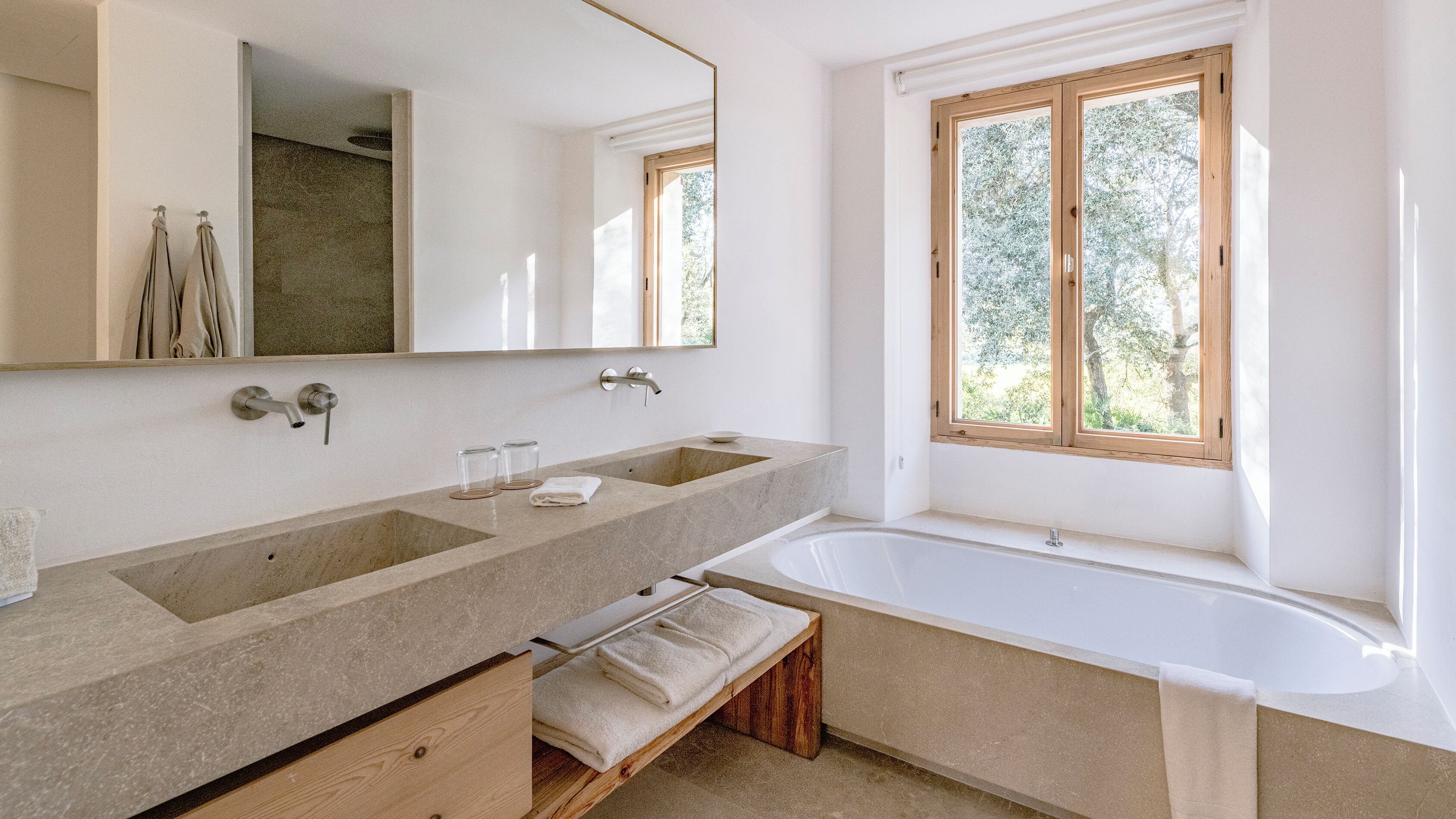 A modern bathroom with two sinks, a large bathtub, a wooden bench, and a window with a view of trees.