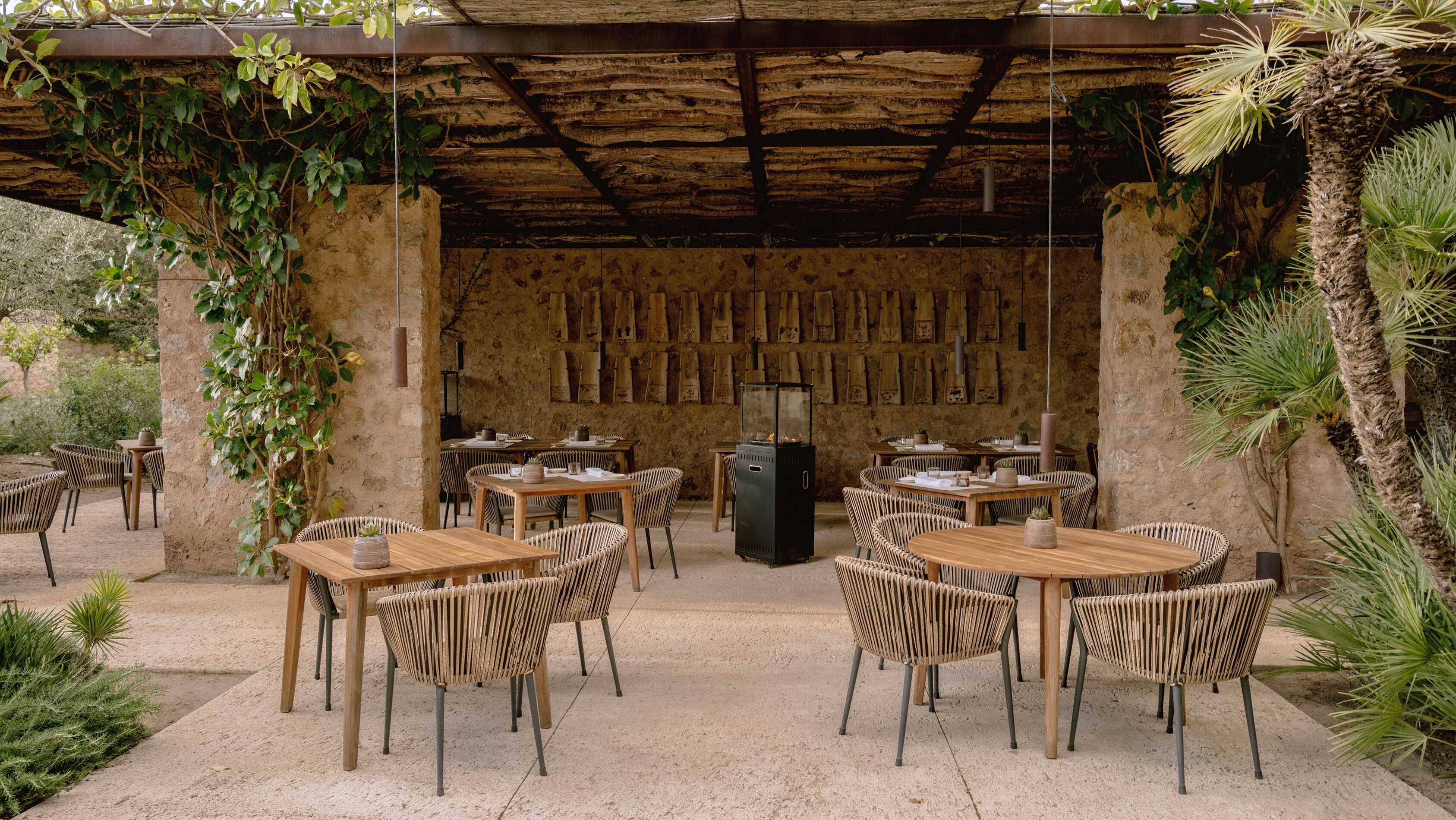Un área de comedor al aire libre con mesas y sillas de madera rústica, rodeada de follaje y un muro de piedra con adornos colgantes.