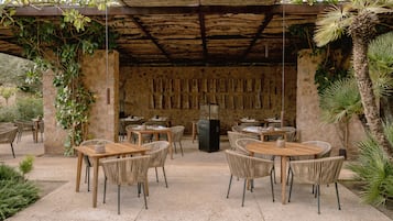Un área de comedor al aire libre con mesas y sillas de madera rústica, rodeada de follaje y un muro de piedra con adornos colgantes.