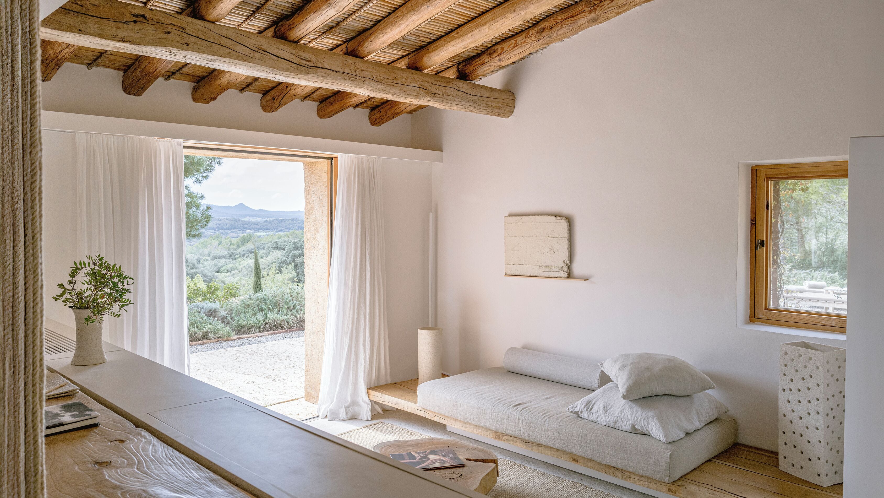 Un dormitorio con techo de madera, una cama con almohadas, una mesita de noche, una ventana con cortinas y vistas al exterior.