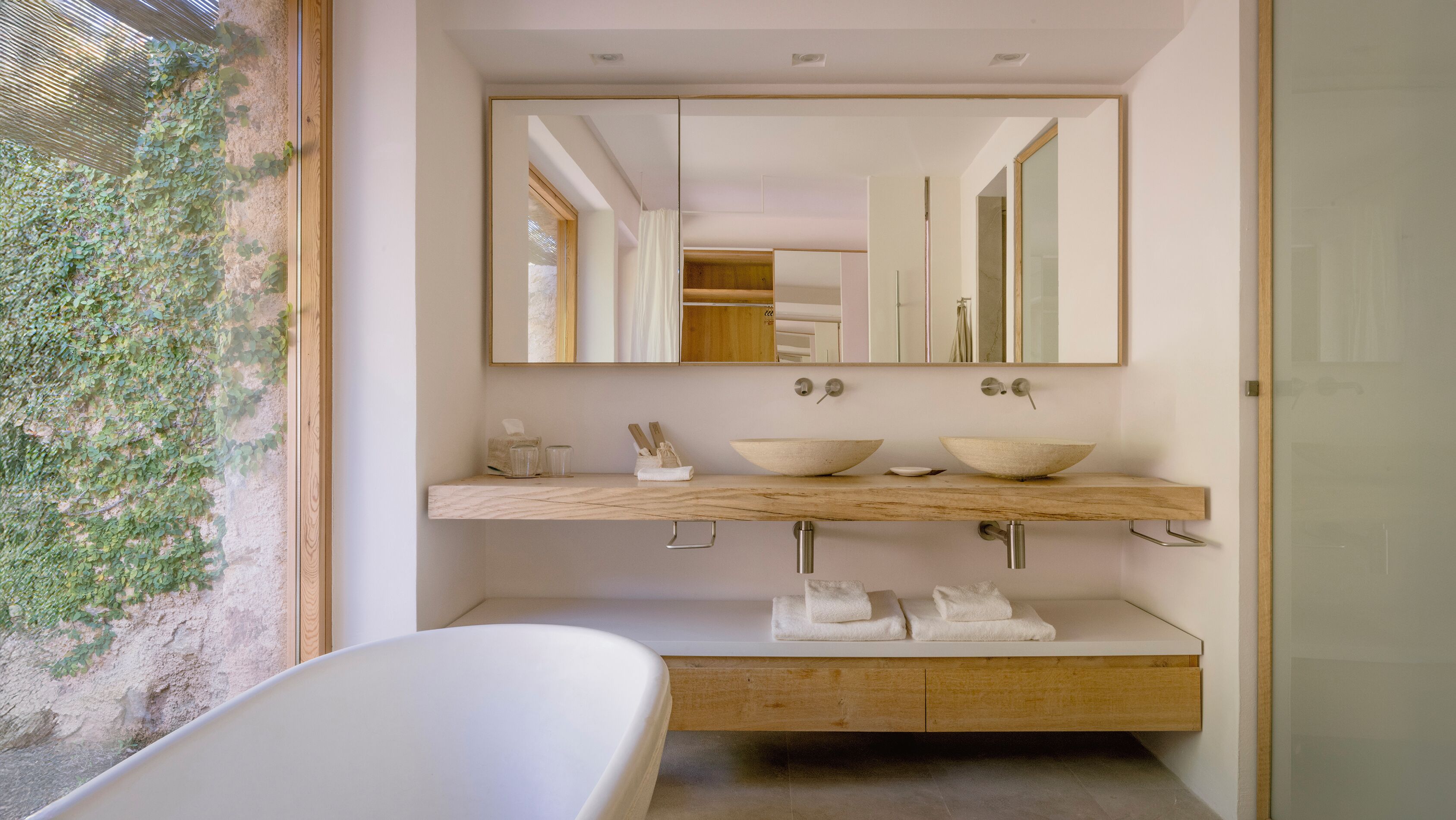 A modern bathroom with a freestanding bathtub, a wooden vanity with two large bowls, a mirror, and a view of a garden through a window.