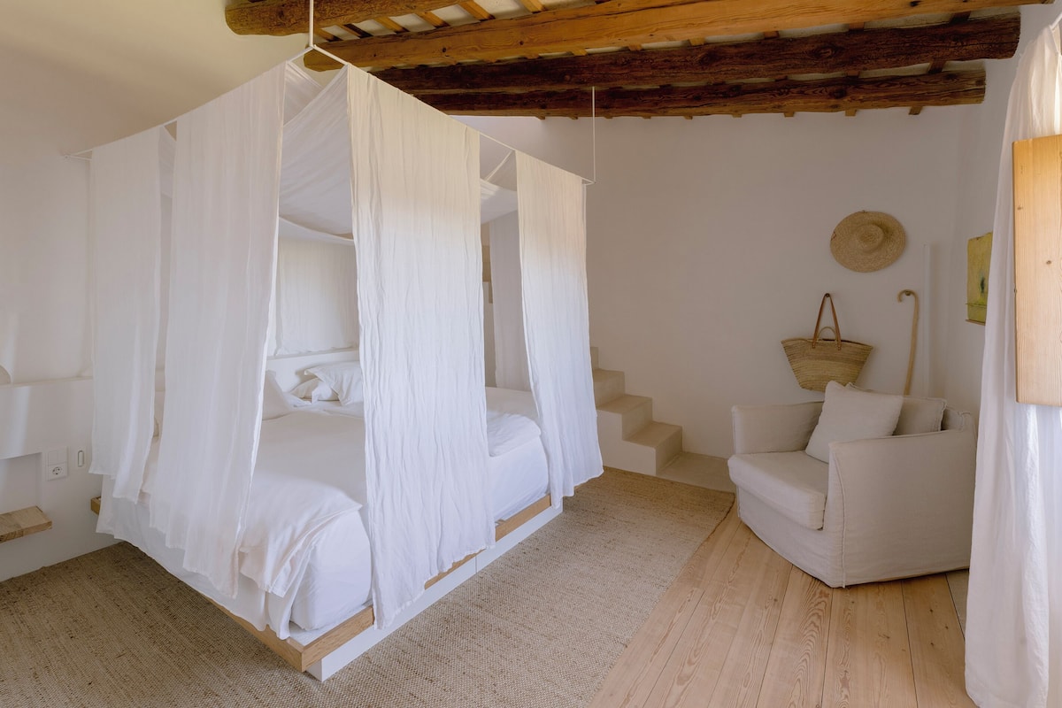 Ein Schlafzimmer mit einem Himmelbett, einem weißen Sessel und einem gewebten Teppich.