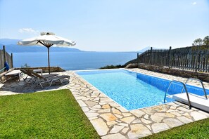 A swimming pool with stone tiles, a grassy area, lounge chairs, and a wooden fence with a view of the sea. - Private Villa with Breathtaking sea view, Swimming pool, Near the beach (Skaloma, Phokis)