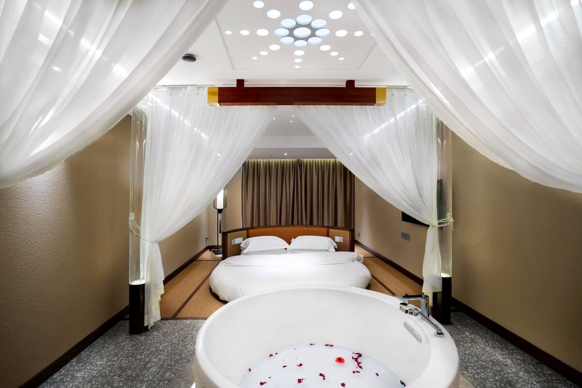 A hotel room with a large bathtub, a bed with white linens, and sheer white drapes.