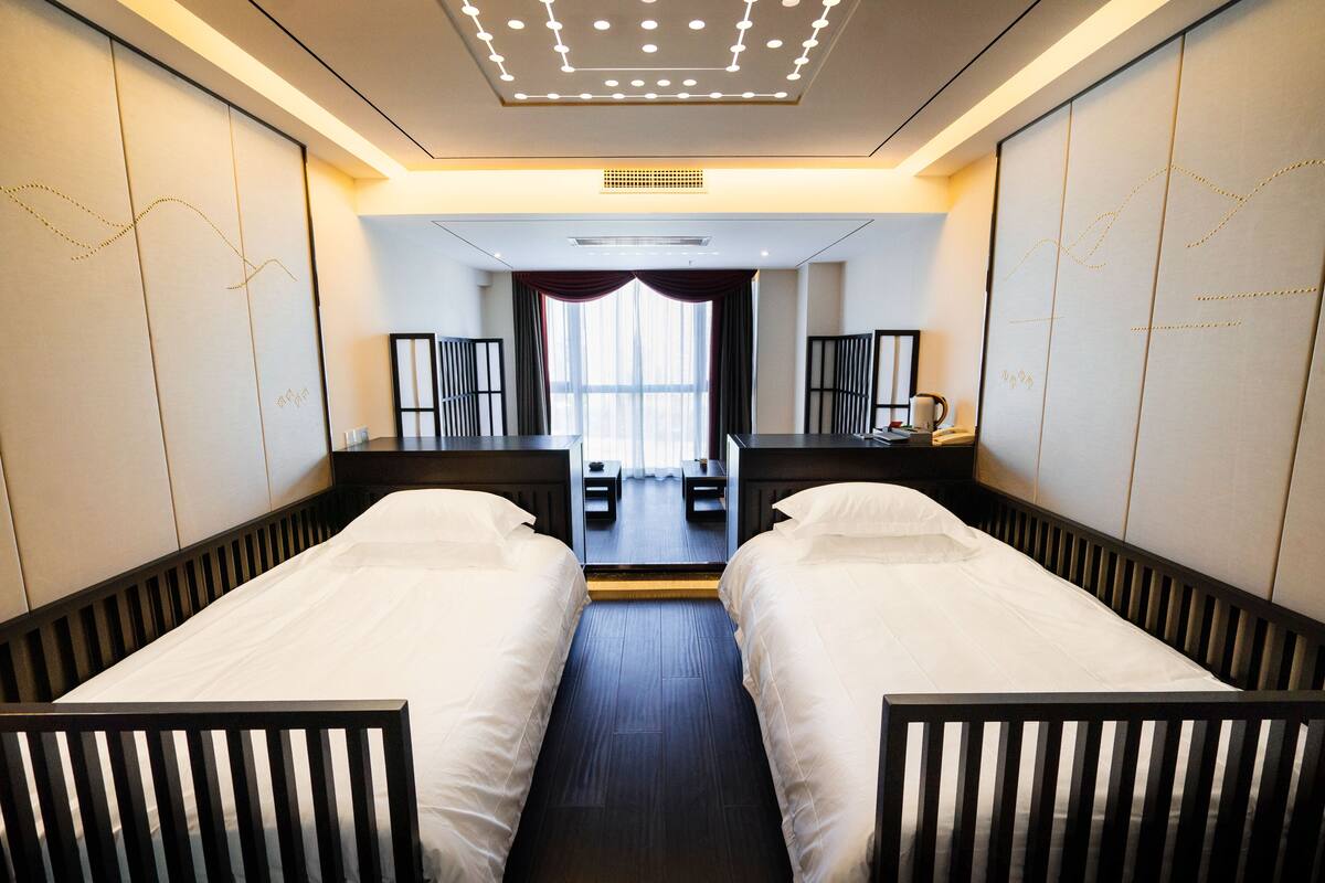 A hotel room with two single beds, a large mirror, a desk with a chair, and a view of the city through large windows.