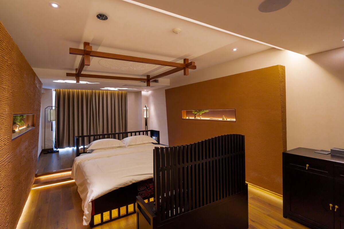 A modern hotel room with a wooden bed frame, a bedside table, a wardrobe, and a view of the cityscape.
