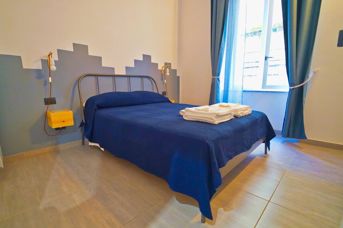 A single bed with a blue bedspread and white towels, a bedside table with a lamp, and a window with blue curtains.