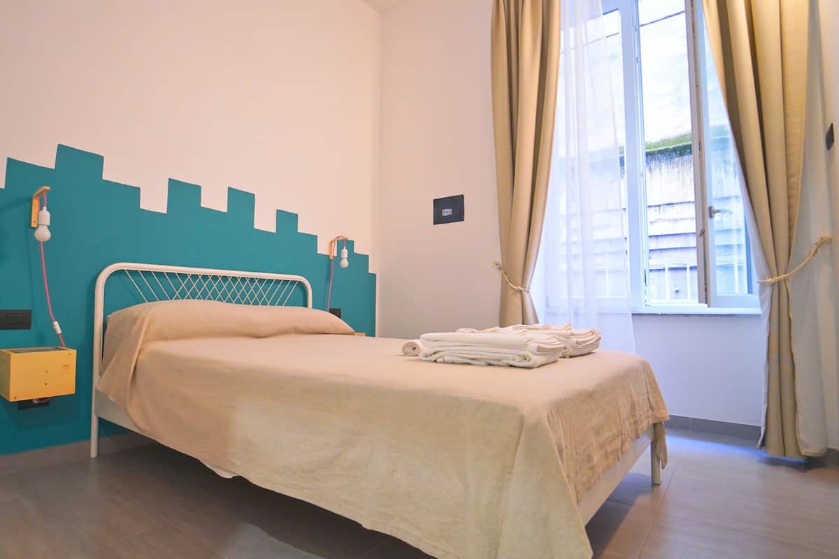 A neatly made bed with a beige bedspread, a bedside table with a lamp, and a window with curtains.