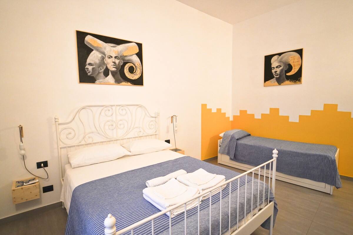 A hotel room with two beds, one larger and one smaller, both with striped bedding. There are two paintings on the wall above the beds, and a small wooden side table with a lamp next to the larger bed.