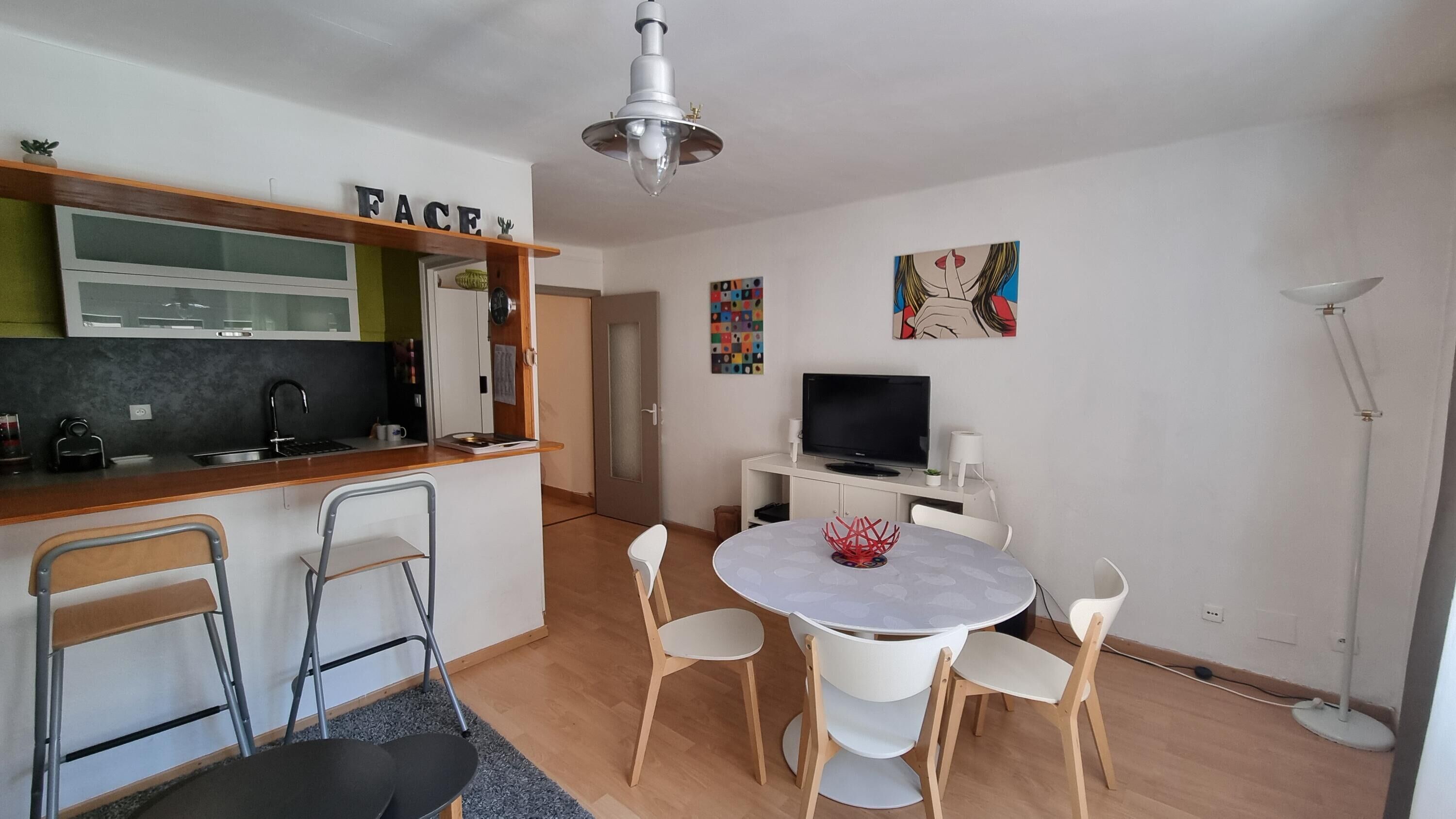 Un espace de vie compact comprenant un coin cuisine, un coin repas avec une table ronde et des chaises, un téléviseur et un luminaire au plafond.