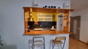 Pieni, käytännöllinen keittiö, jossa on puinen työtaso, kaksi metallituolia, pesuallas ja seinällä oleva kello.
