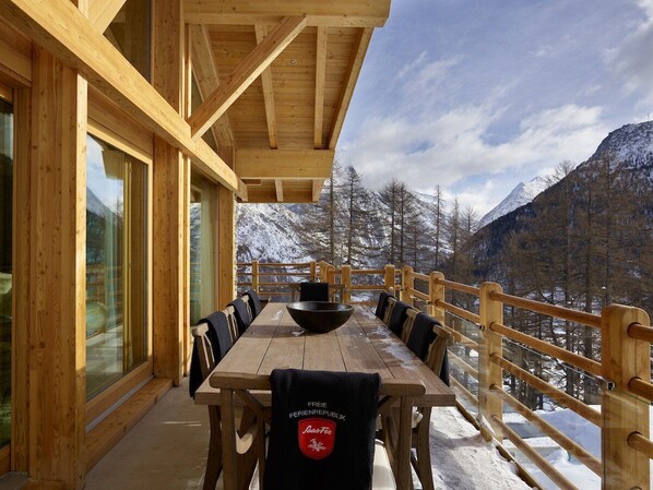 A wooden deck with a dining table and chairs, overlooking a snowy mountain landscape.